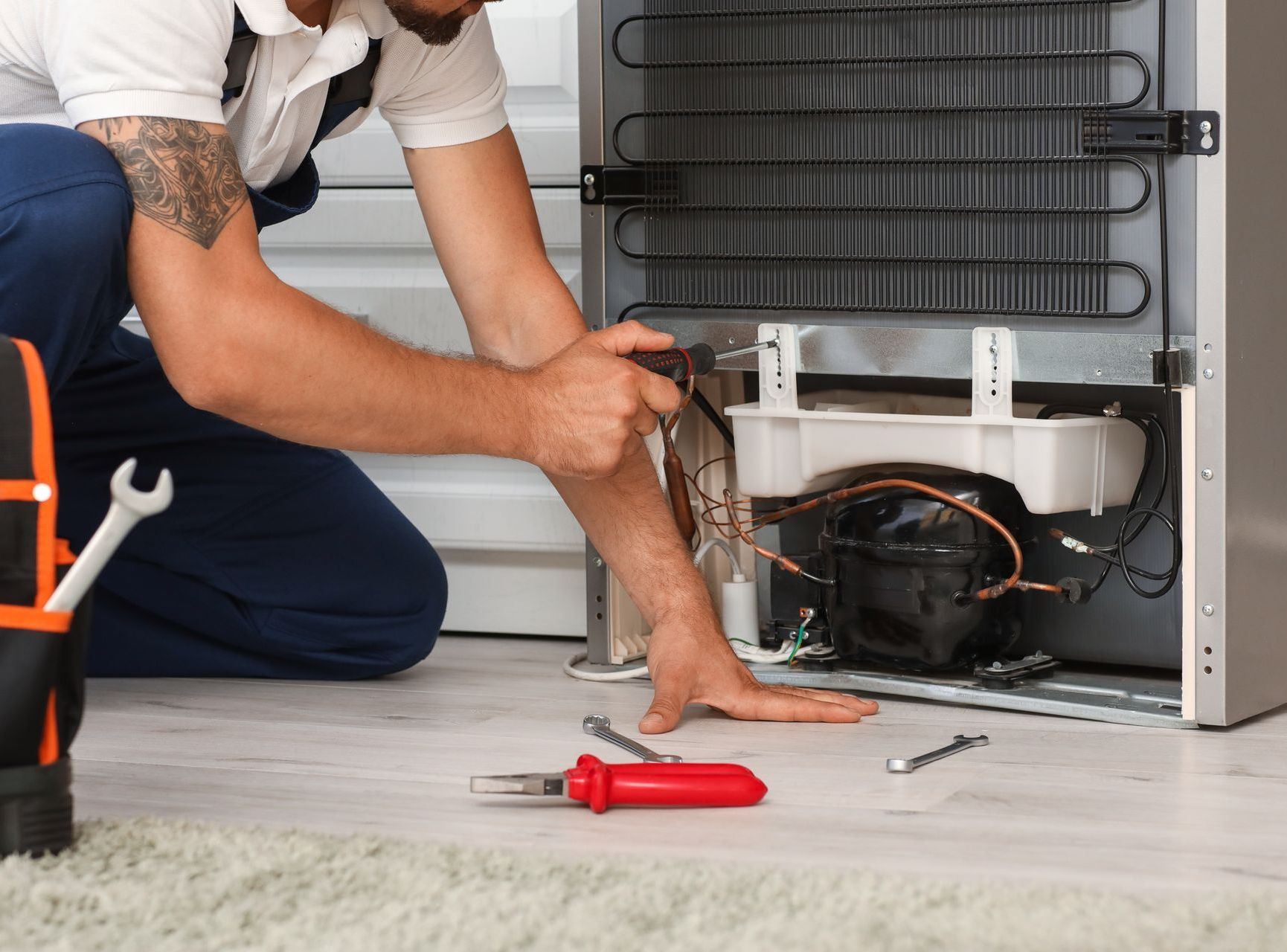 A man is kneeling on the floor fixing a refrigerator.
