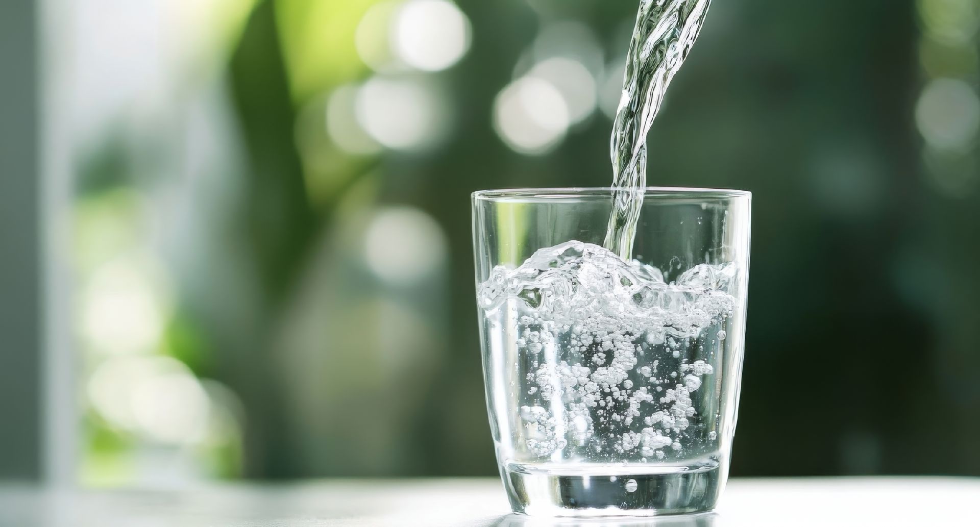 Water being poured into a clear glass on a bright table, with a blurred green background.