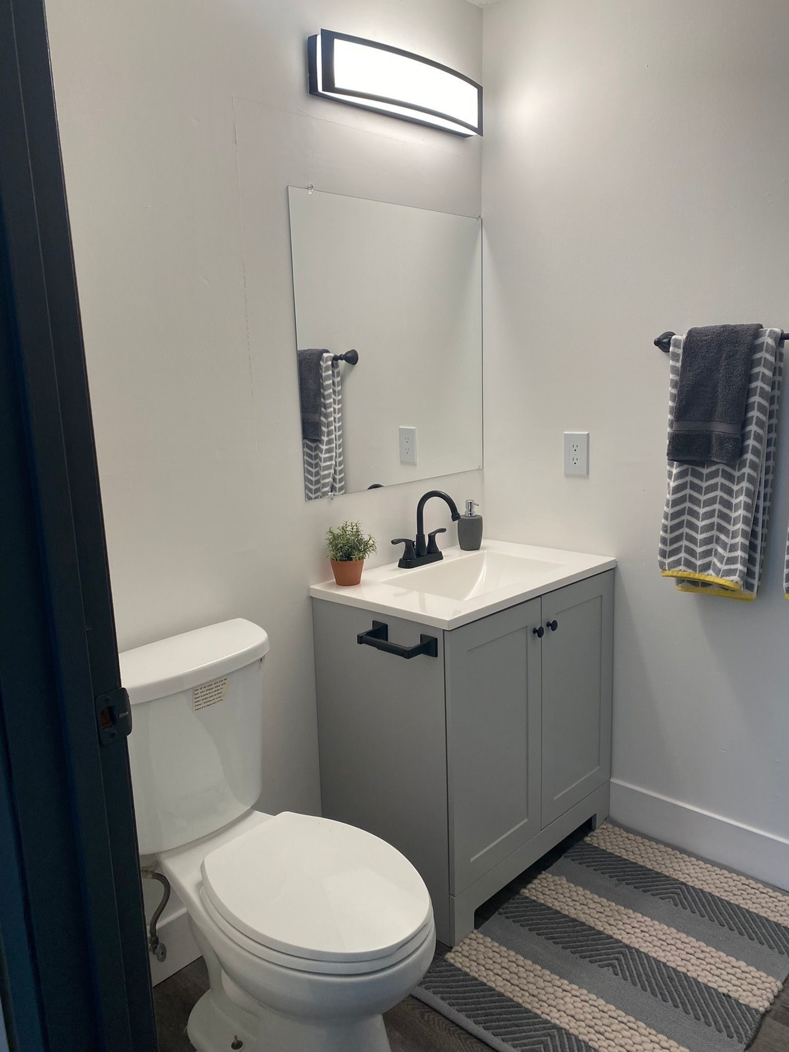 Small bathroom with gray vanity, white sink, black faucet, toilet, towel rack, and striped rug.