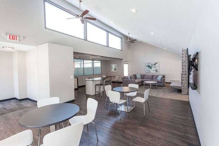 Open community lounge with round tables, white chairs, a gray sectional sofa, ceiling fans, and a wall-mounted TV.