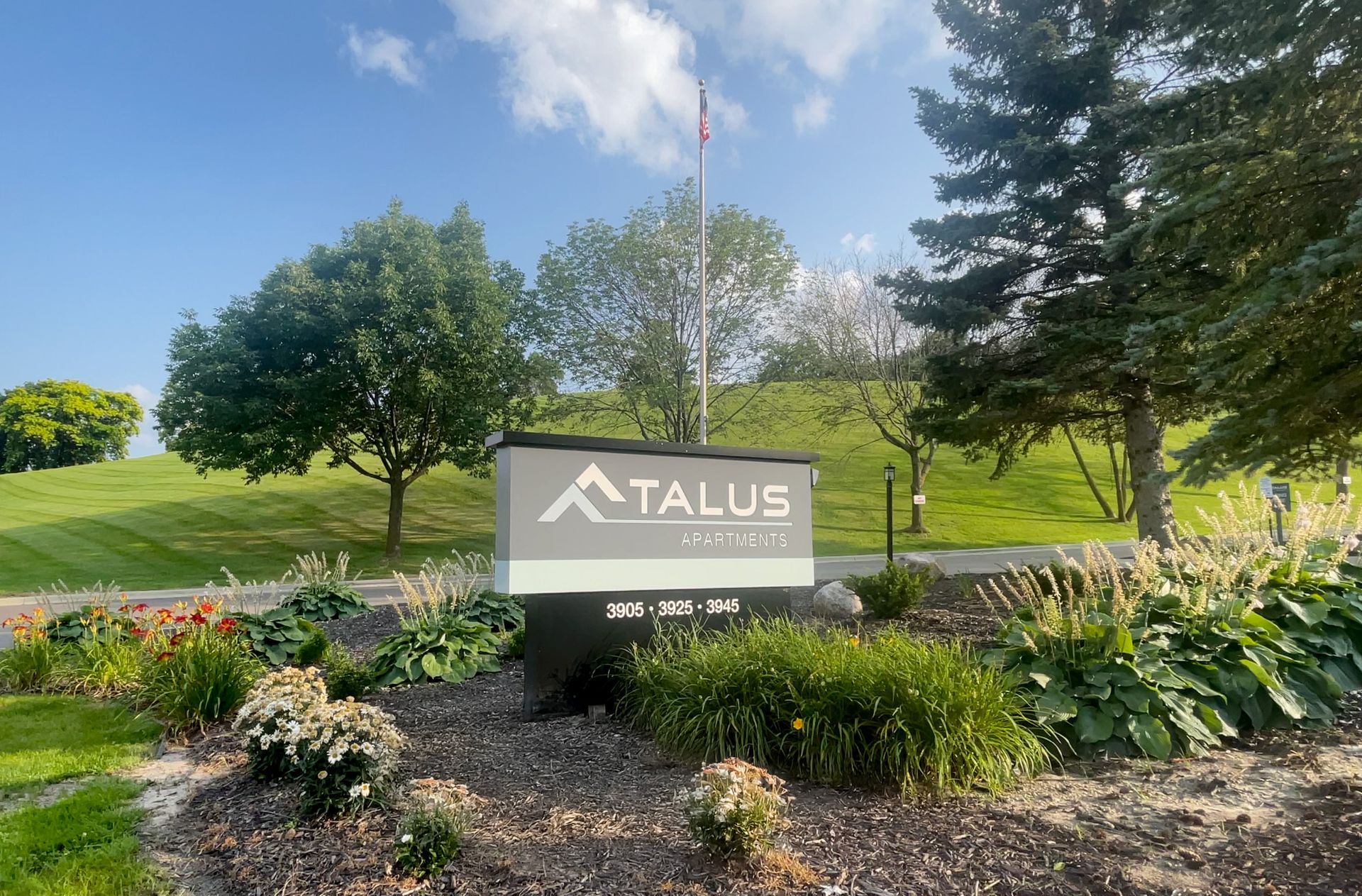 Talus Apartments sign in landscaped garden with trees and blue sky.