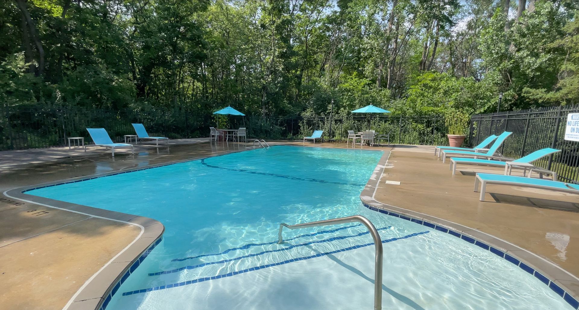 Outdoor swimming pool with blue lounge chairs and umbrellas surrounded by trees.