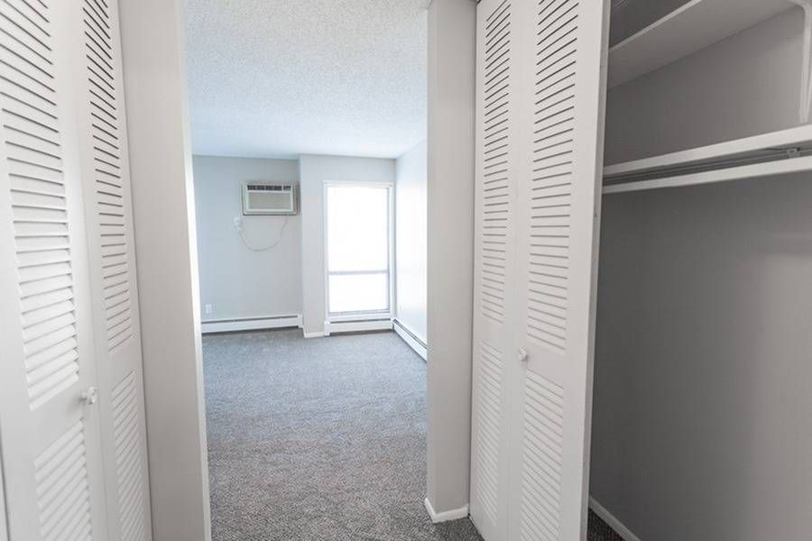 Bedroom with white louvered closet doors on both sides, carpet, a window, and a wall-mounted air conditioner.