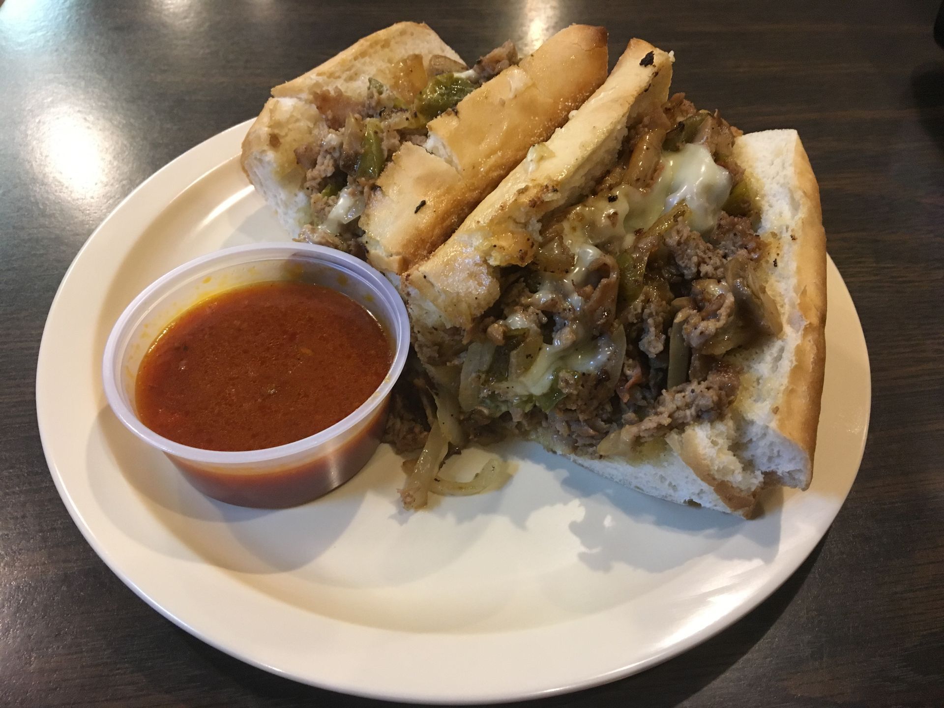 A steak and cheese sub sandwich with a side of sauce on a white plate. A steak and cheese sub sandwich with a side of sauce on a white plate.