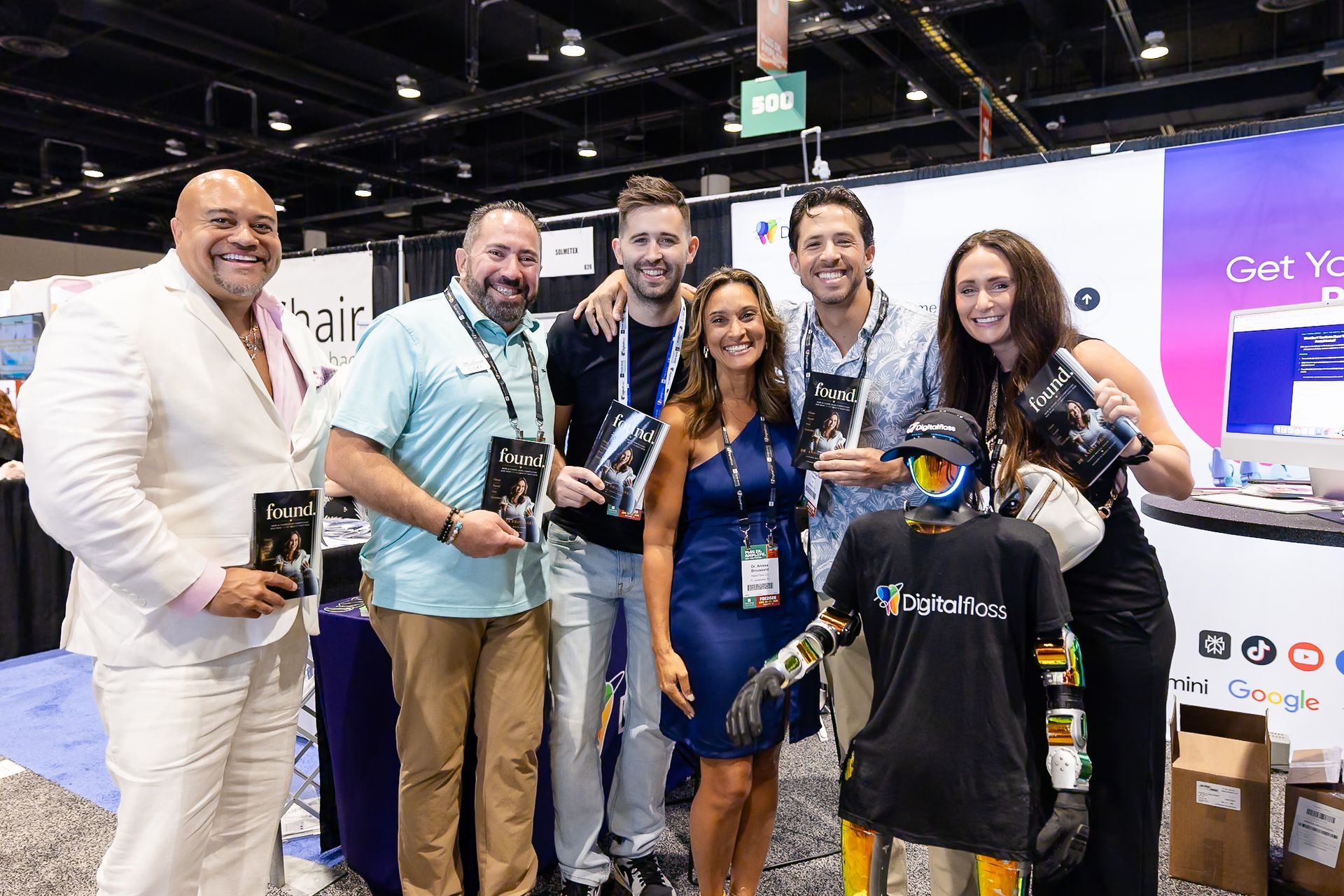 Group of people posing at a trade show booth, holding cameras and smiling in a convention hall.