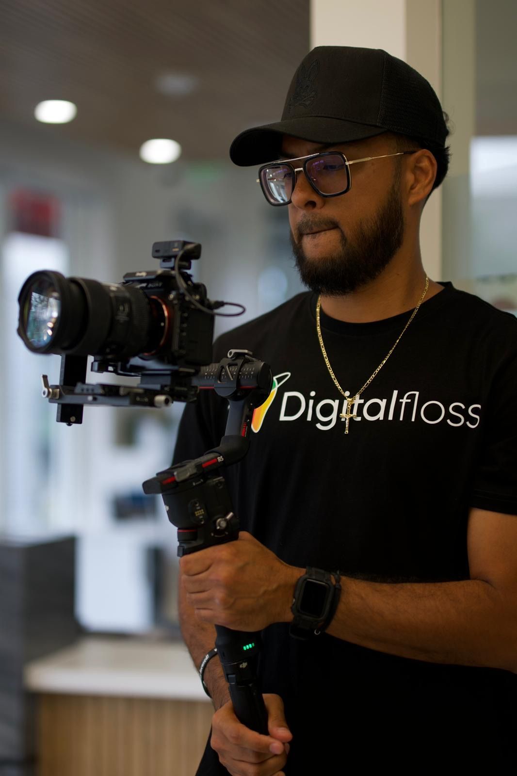 Man in black hat, glasses, and t-shirt with camera on a stabilizer, indoors.