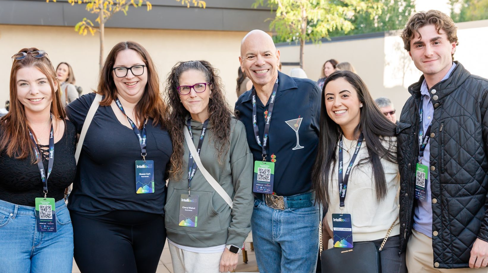 Group of people at an event, smiling and posing. Some have lanyards. Indoors, bright lighting.
