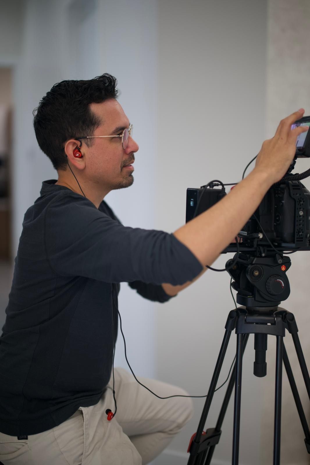 Man with glasses, adjusting camera on tripod in front of a white wall. He's wearing earbuds and light pants.