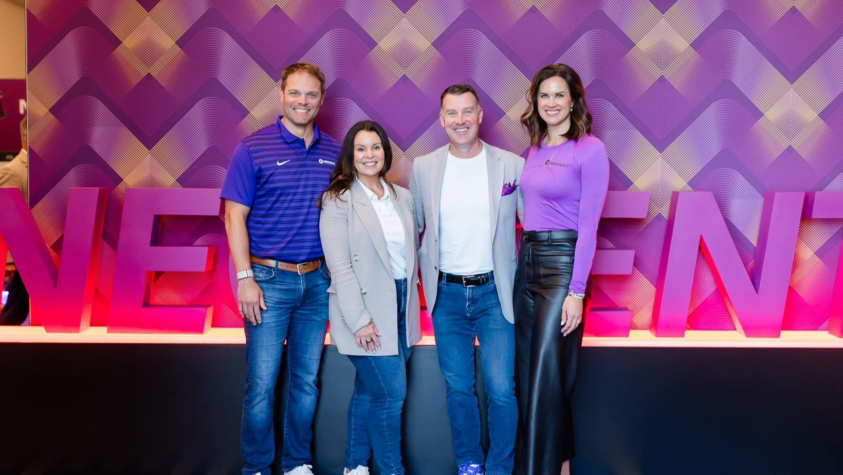 Group of people at an event, smiling and posing. Some have lanyards. Indoors, bright lighting.