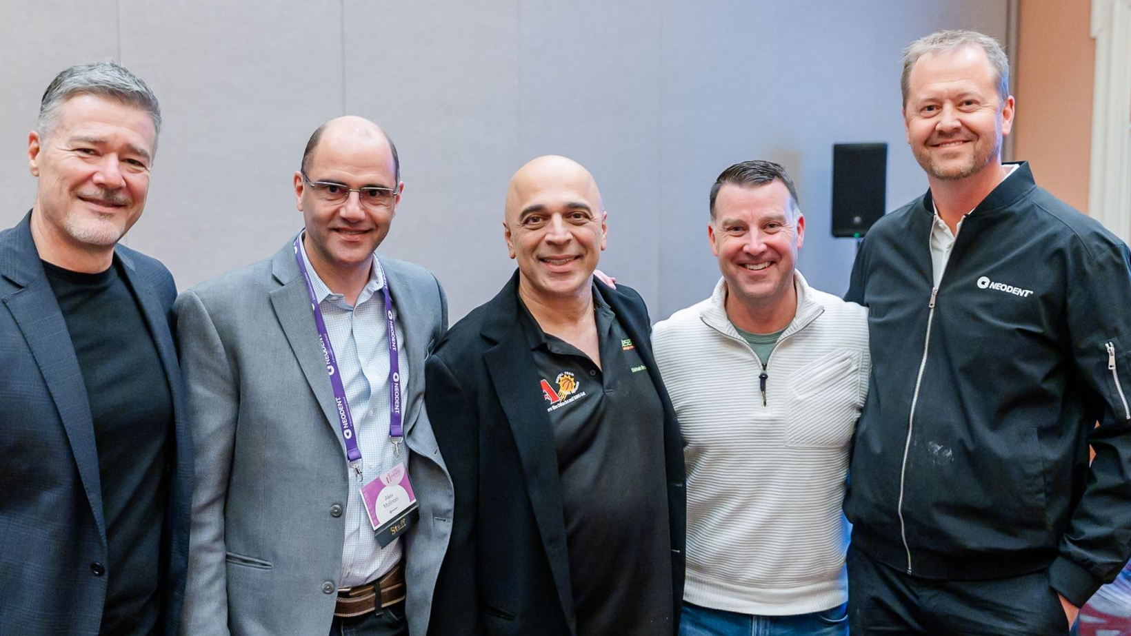 Group of people at an event, smiling and posing. Some have lanyards. Indoors, bright lighting.