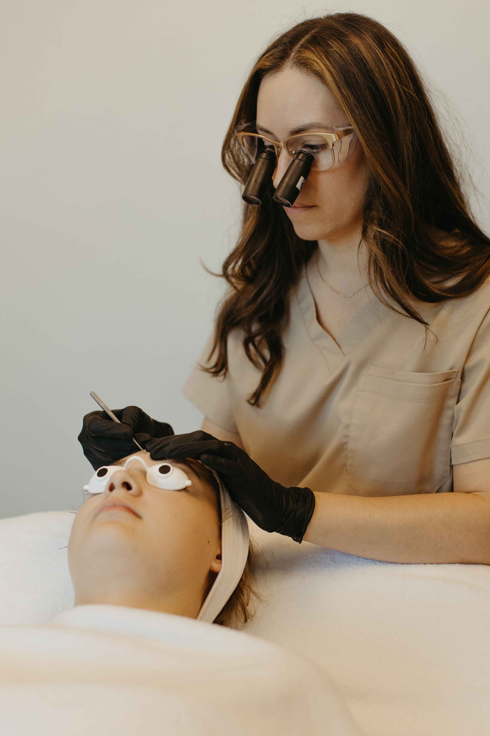 A woman with magnifying glasses, and black gloves performing a cosmetic procedure on a patient.