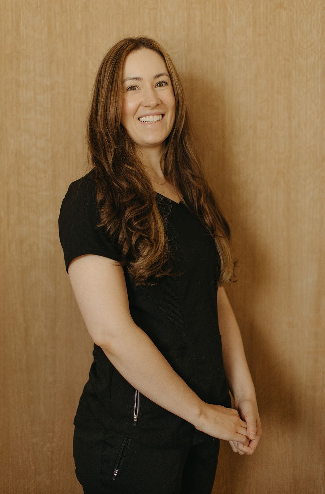 Woman with long brown hair smiling, wearing a black top and pants, standing in front of a wood-paneled wall.