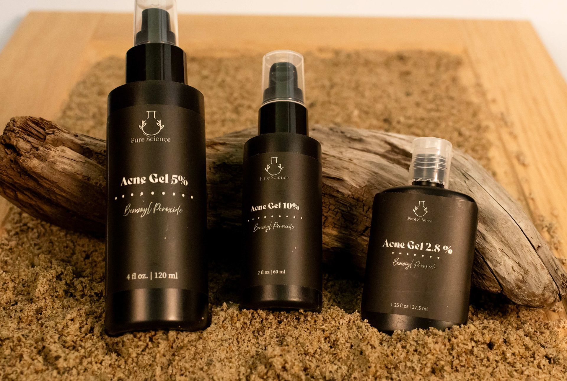 Three dark brown cosmetic bottles with white labels, arranged on sand and driftwood.