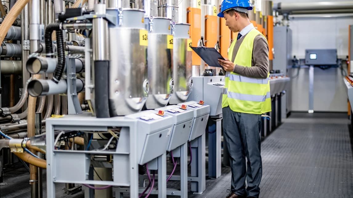 A man is standing in a factory looking at a clipboard.