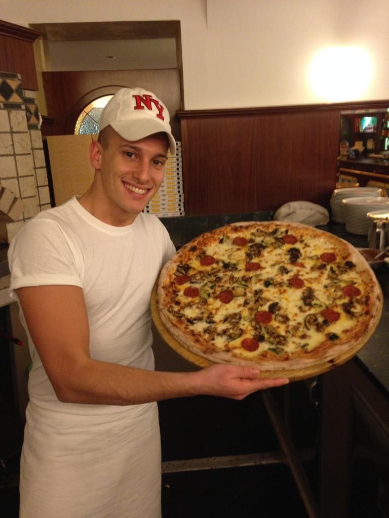 Uomo con camicia bianca e cappellino che tiene in mano una pizza grande nella cucina di un ristorante