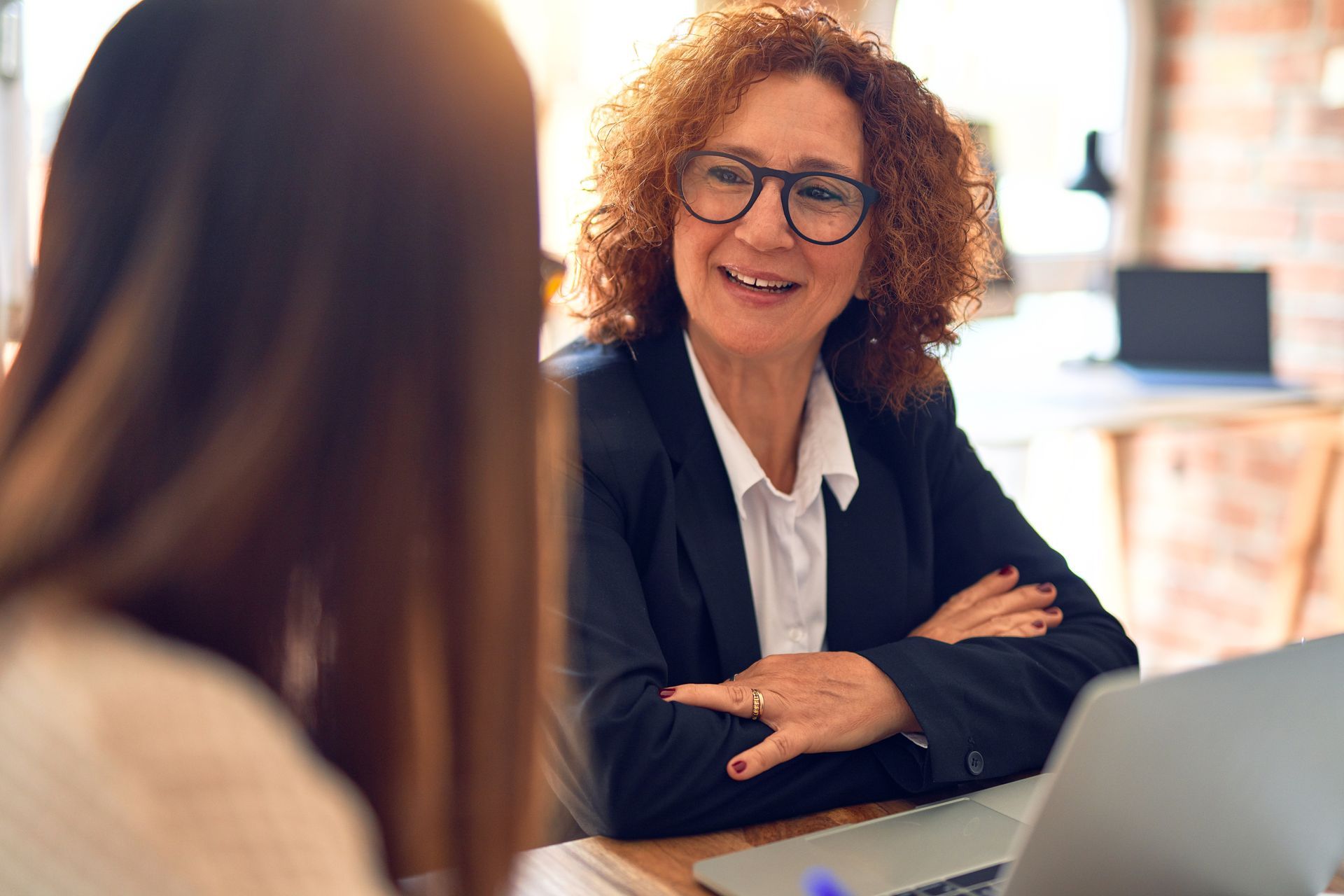 A person with curly hair and glasses smiles while talking to another person in a workspace. A person with curly hair and glasses smiles while talking to another person in a workspace.