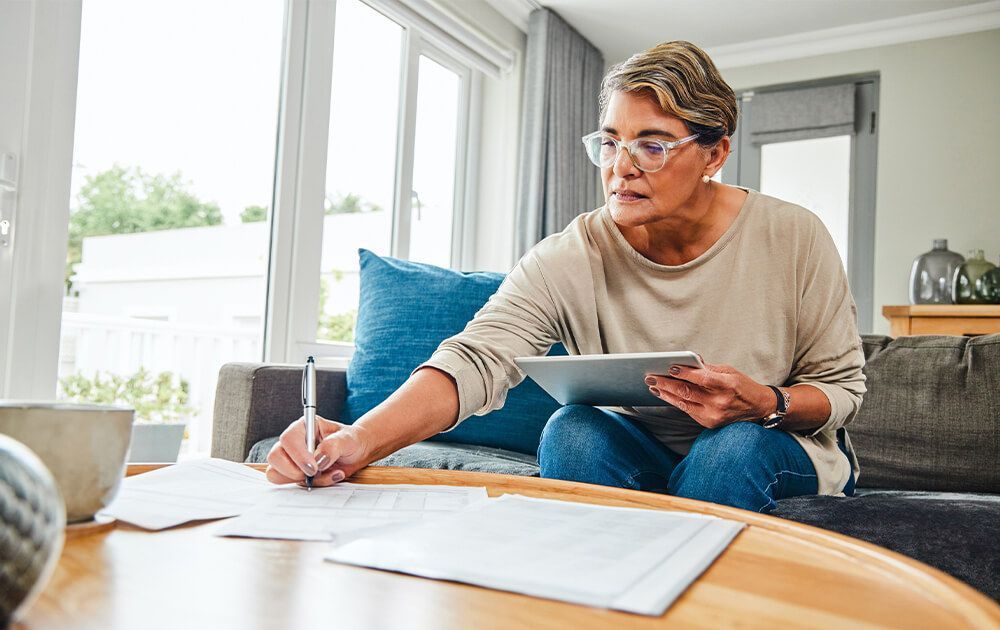 a woman sits on a couch writing on a piece of paper while using a tablet