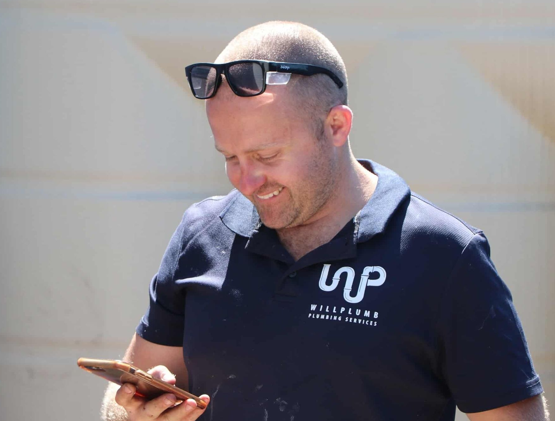 A person wearing dark sunglasses and a navy polo shirt with a logo, smiling while looking down at a handheld device.