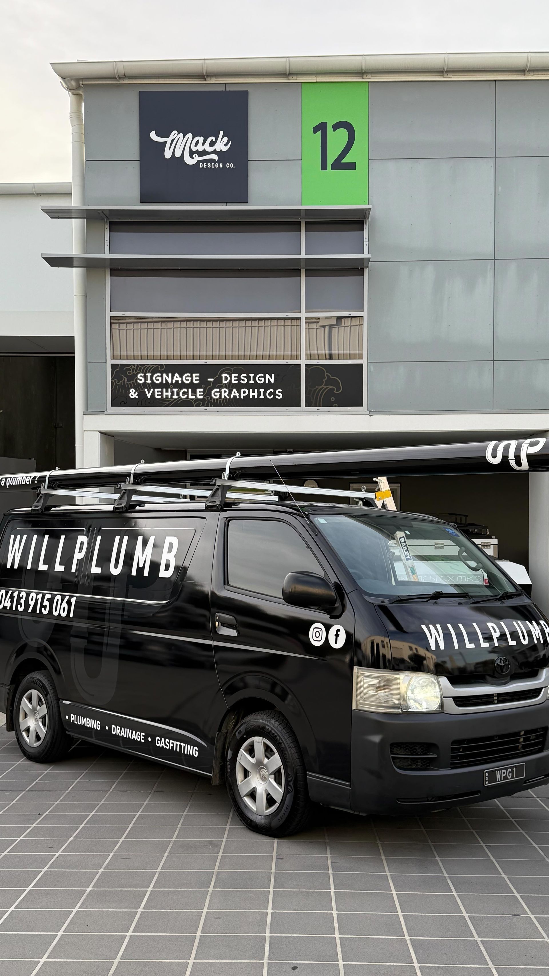 A black Willplumb work van parked in front of a commercial building labeled unit 12.