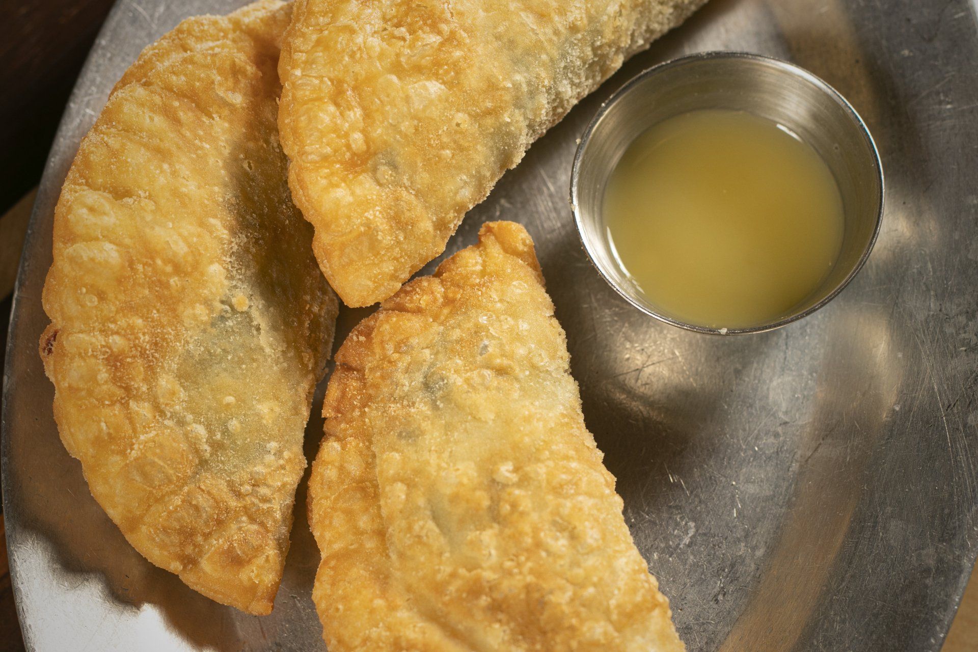 Three golden-brown, fried pastries with a small bowl of yellow dipping sauce on a metal plate.
