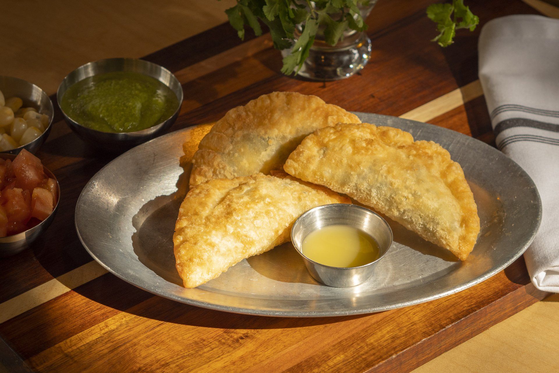 Golden fried pastries on a metal plate with dipping sauce and various chutneys on a wooden surface.