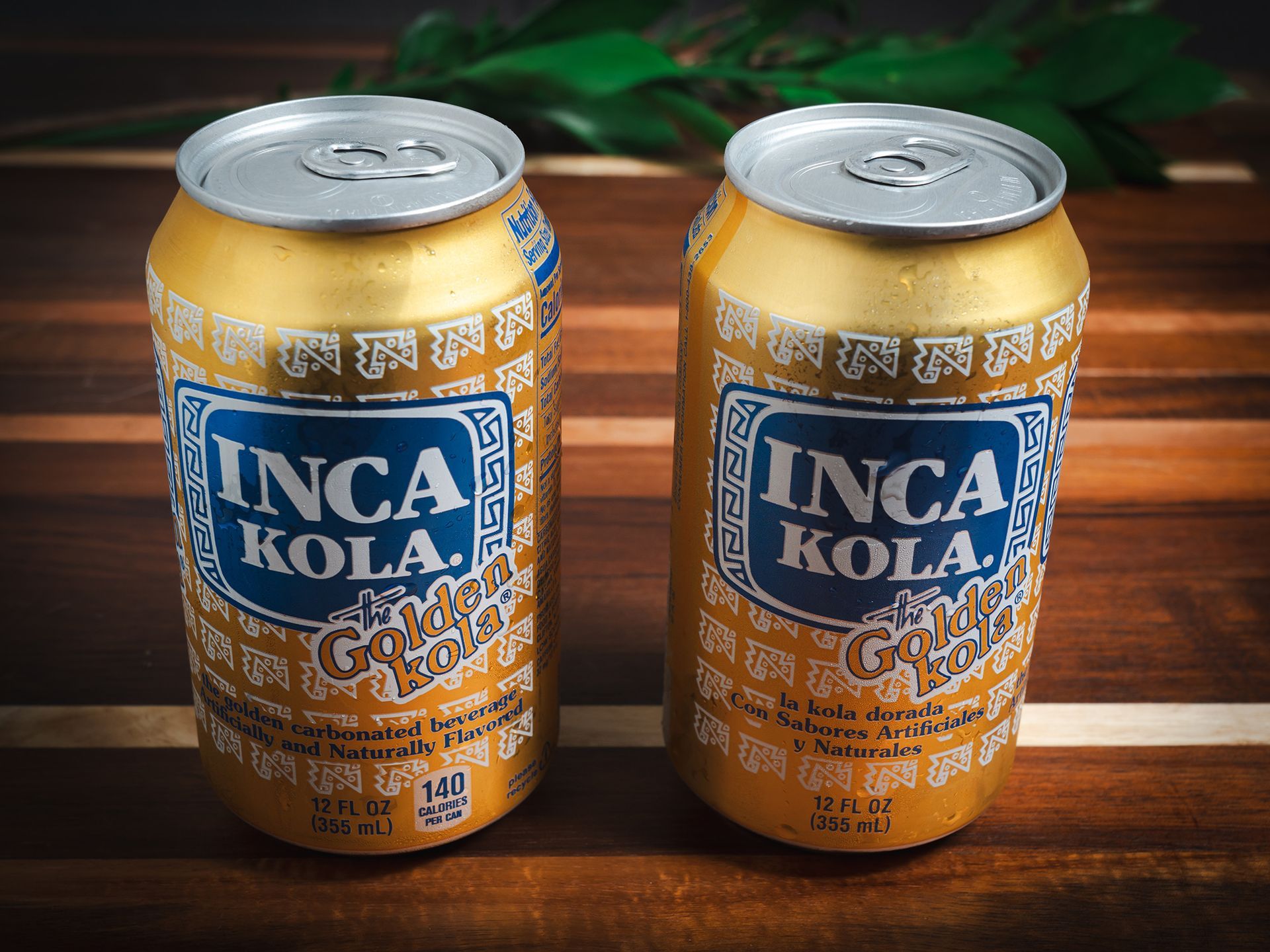 Two cans of Inca Kola, a golden-colored soda, on a wooden surface.