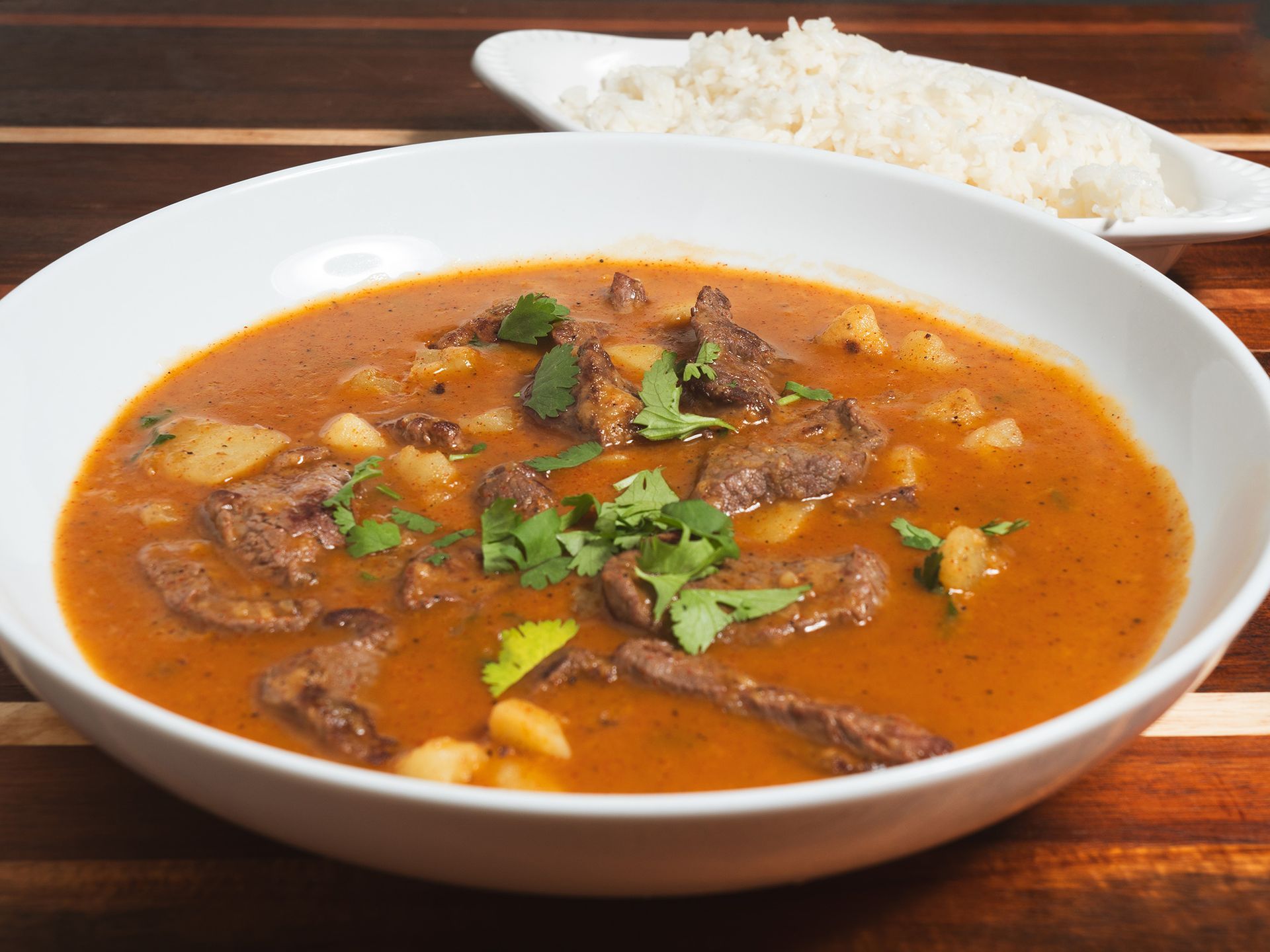 Beef stew with potatoes and cilantro in a white bowl, served with white rice on a wooden surface.