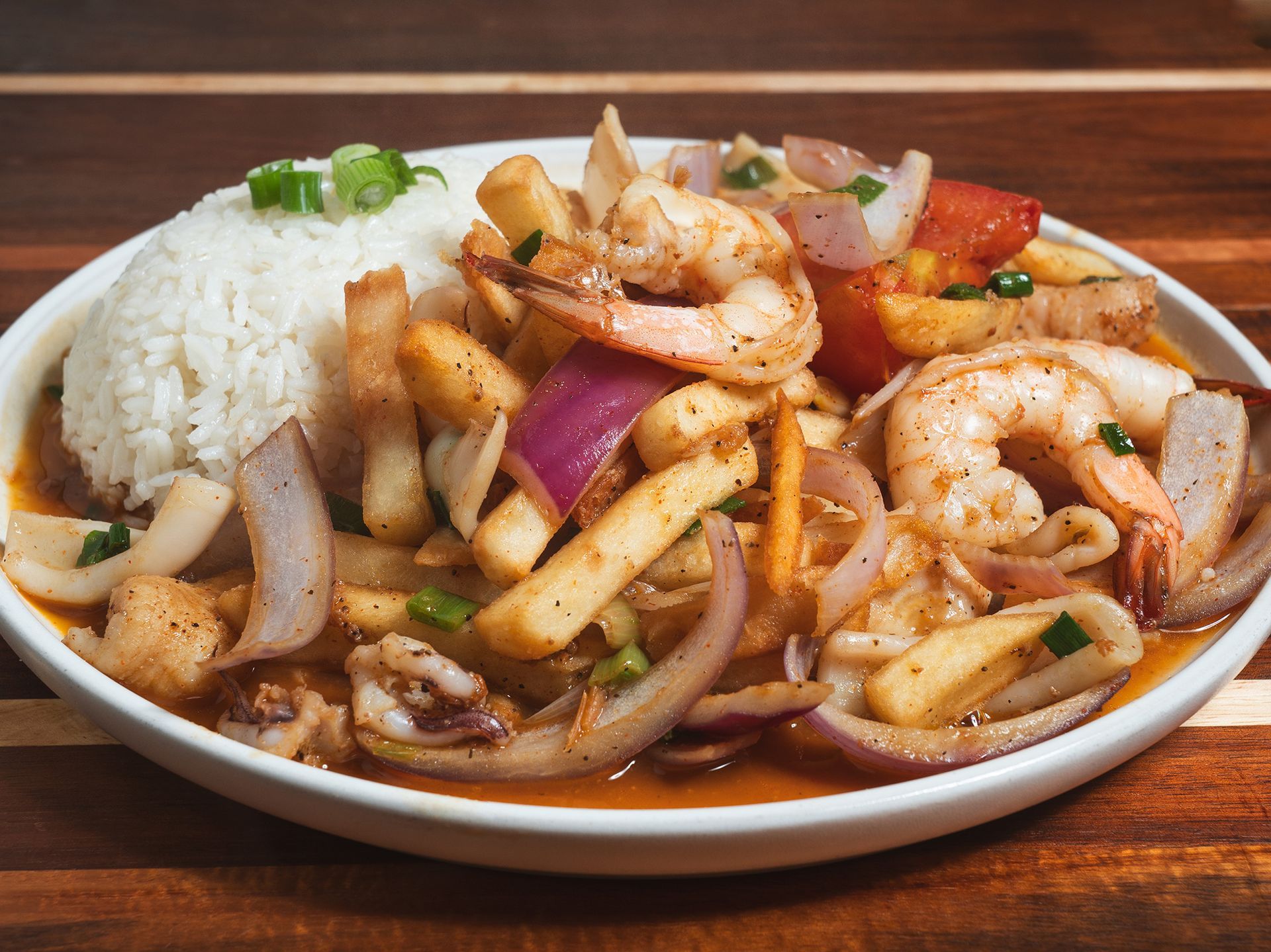 Plate of Peruvian-style seafood stir-fry with rice, including shrimp, squid, onions, tomatoes, and fries.