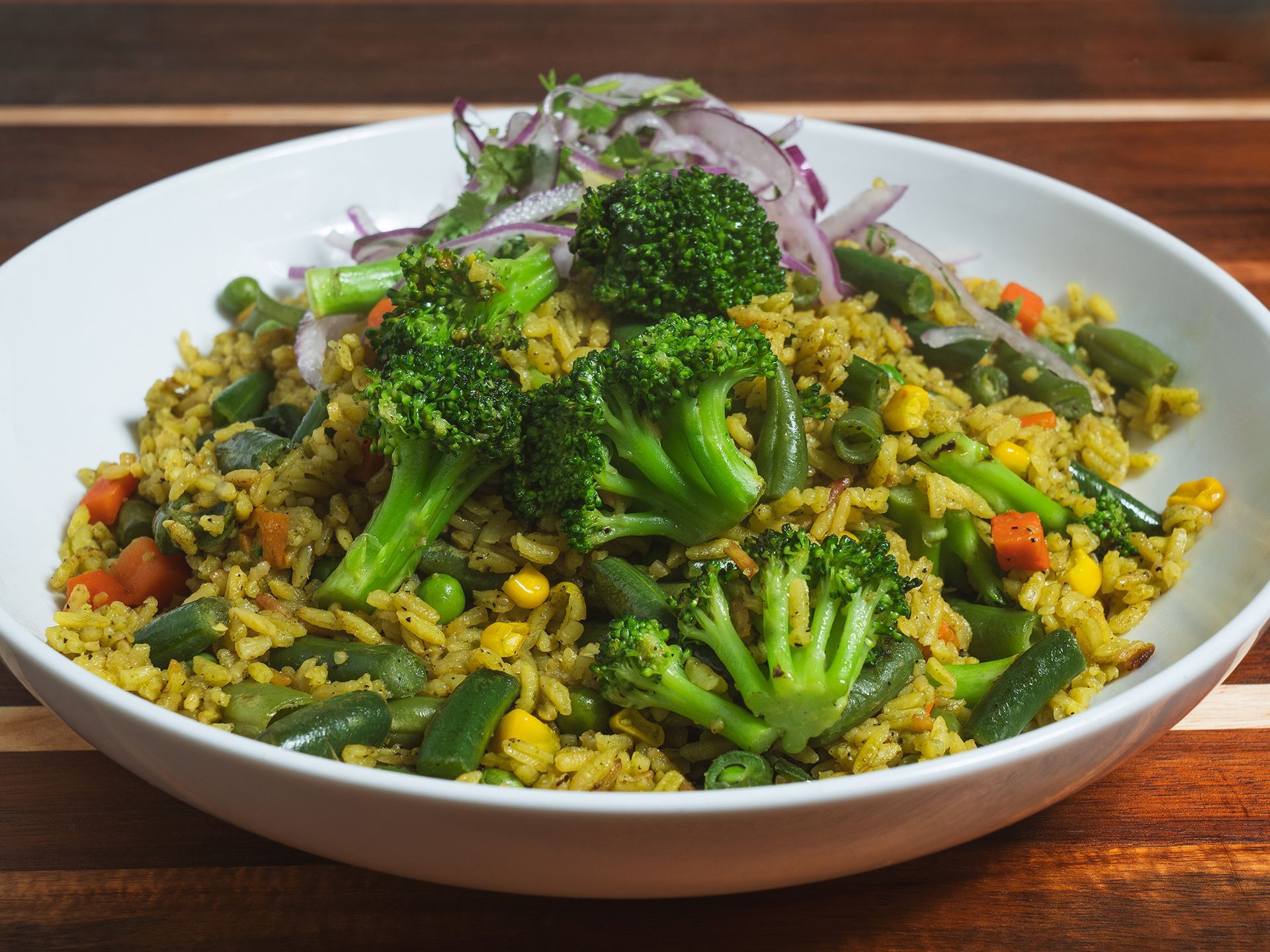 Vegetable fried rice in a white bowl, topped with broccoli florets and red onion, on a wooden surface.