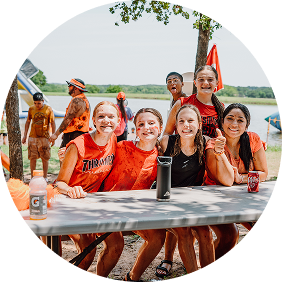 Group of friends in orange shirts smiling at an outdoor picnic table by a lake, holding drinks under a tree