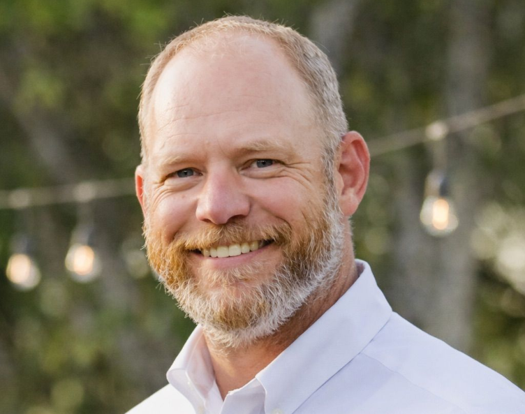 Smiling man with a beard in a white shirt standing outdoors with blurred trees behind him