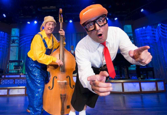 Two performers on a blue-lit stage, one playing upright bass and another leaning toward the camera in glasses and orange beret
