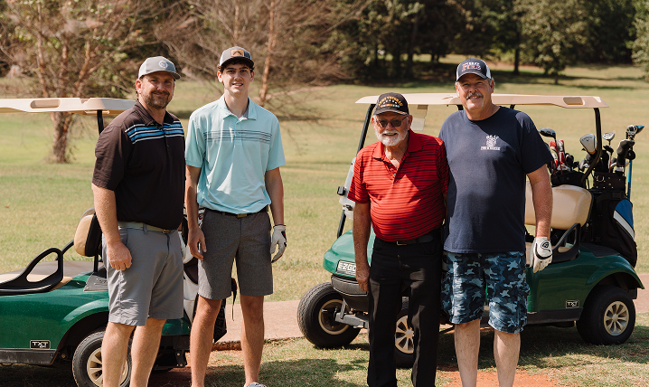 Four people standing beside golf carts on a sunny course, smiling at the camera