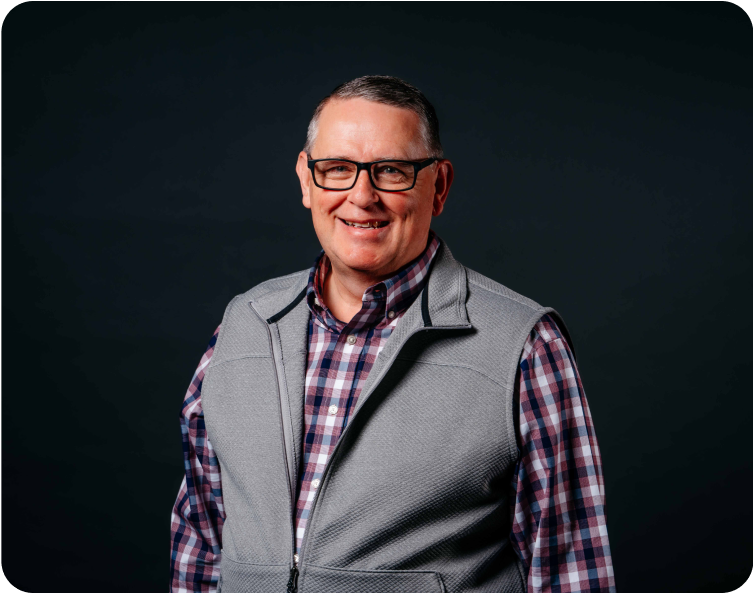 Smiling person in glasses wearing a plaid shirt and gray vest against a dark background