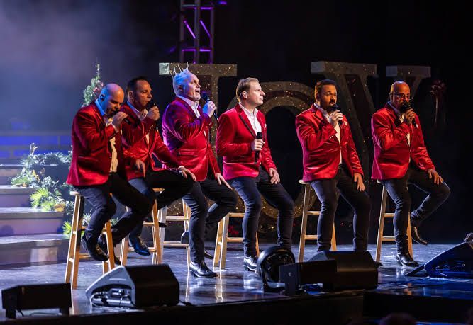 Six singers in red jackets performing on a stage with microphones and stools