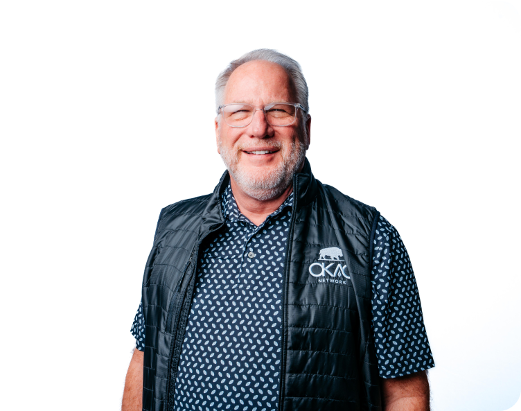 Smiling man wearing glasses, a navy patterned shirt, and black vest against a white background