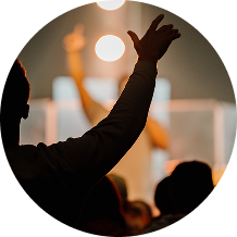 Silhouetted raised arms in a crowd, with blurred stage lights and a speaker in the background