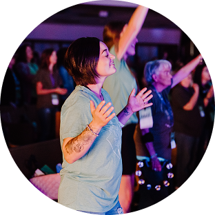 Woman with short dark hair in a light blue shirt, singing and raising hands at a lively indoor event
