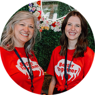 Two smiling women in red “Better Together” shirts at a floral event backdrop