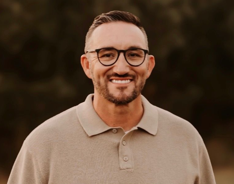 Smiling man with glasses in a beige polo shirt against a blurred brown background