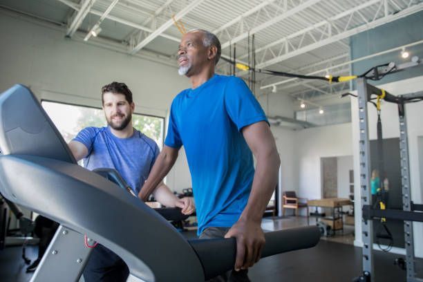 Man walking on treadmill with trainer's assistance in a gym. Blue shirts, indoor.