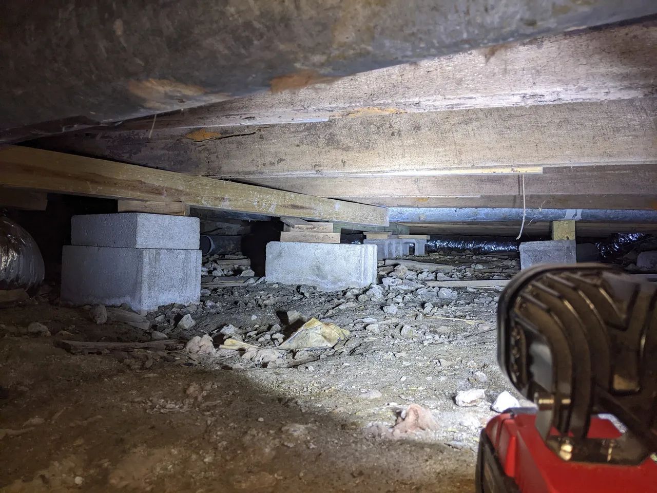 Low crawl space with concrete blocks supporting floor joists and a flashlight in the foreground