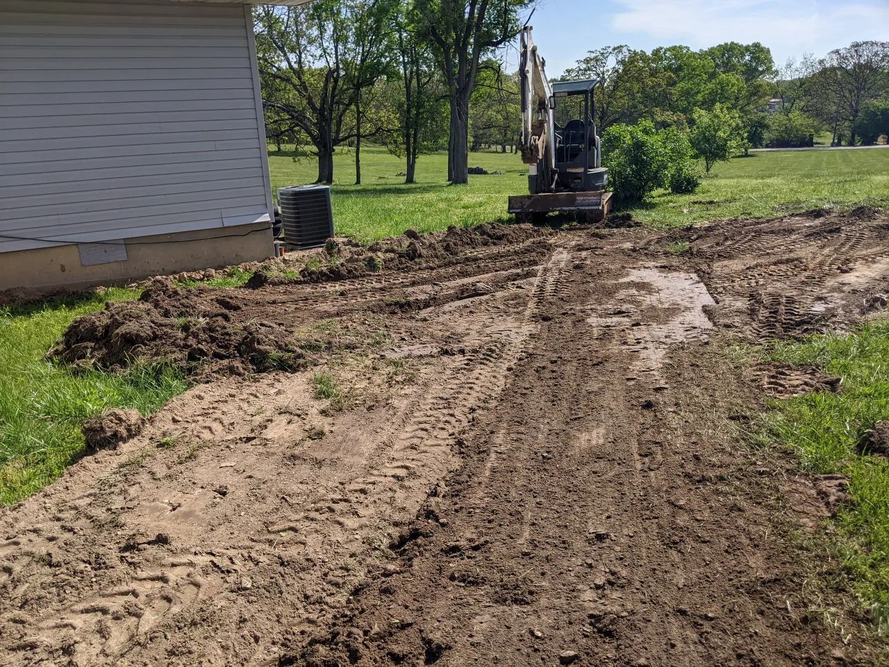 Backyard excavation beside a white house, with a small excavator and muddy dirt path