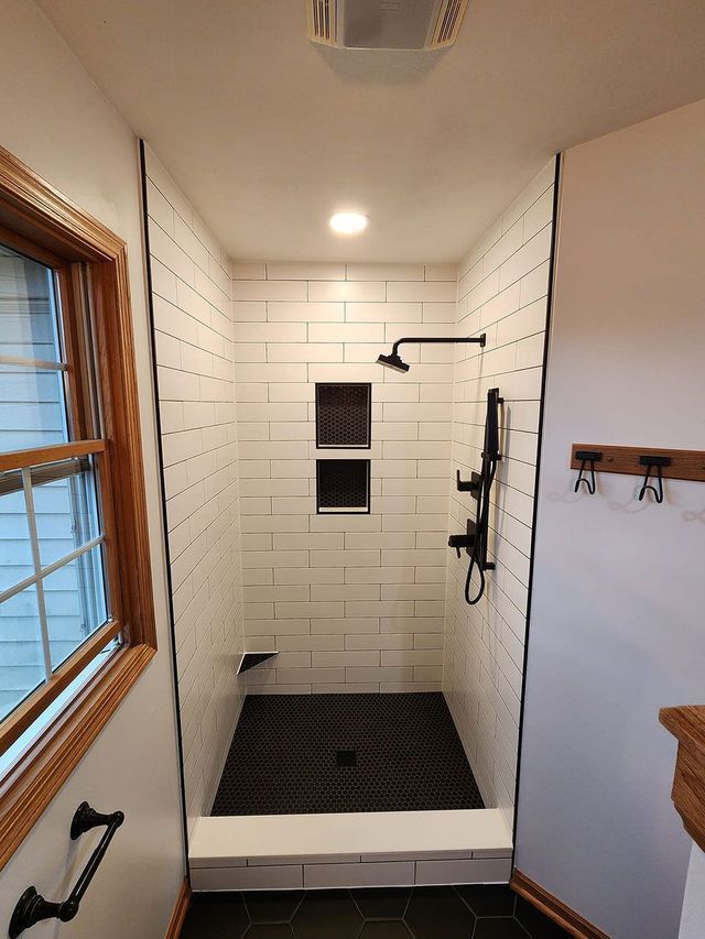 Narrow tiled shower with black fixtures, recessed shelf, and bench, viewed from the bathroom doorway