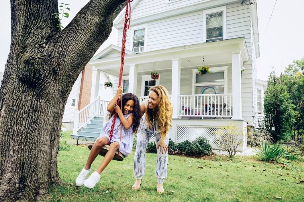 Two children on a rope swing in a yard, smiling beside a white house with a large tree.