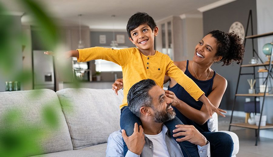 Smiling family playing indoors, child on father's shoulders beside a woman in a bright living room