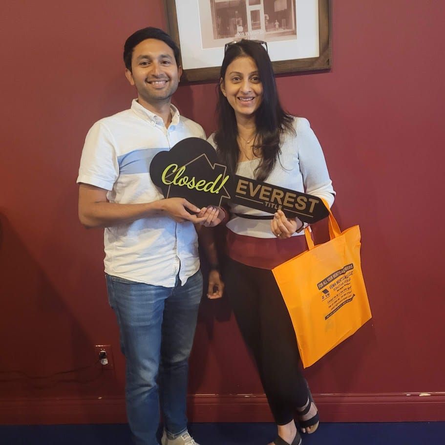 Two people smiling and holding a Closed Everest plaque, standing against a maroon wall with an orange shopping bag.