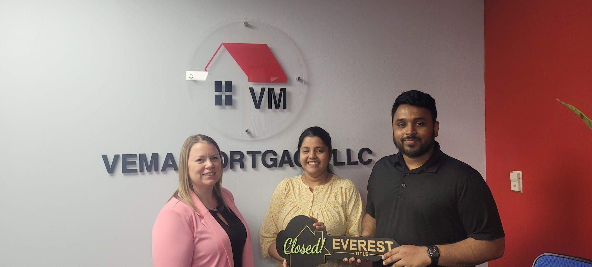 Three people pose indoors holding a “Great Everest” sign in front of a VEMA Mortgage LLC wall logo.