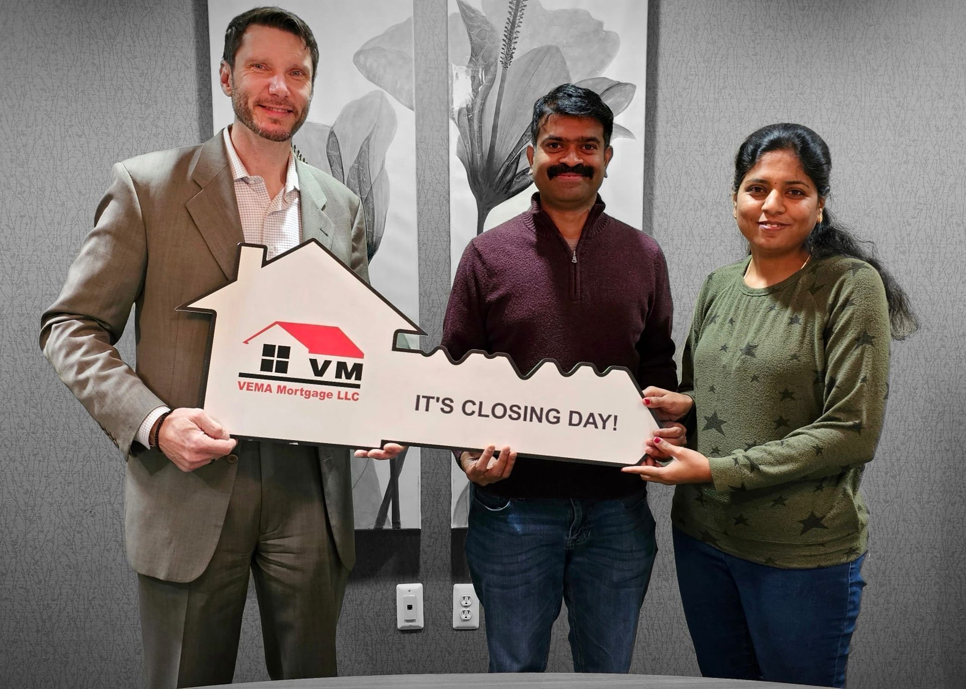 Three people hold a house-shaped sign reading “IT’S CLOSING DAY” in front of a mural.