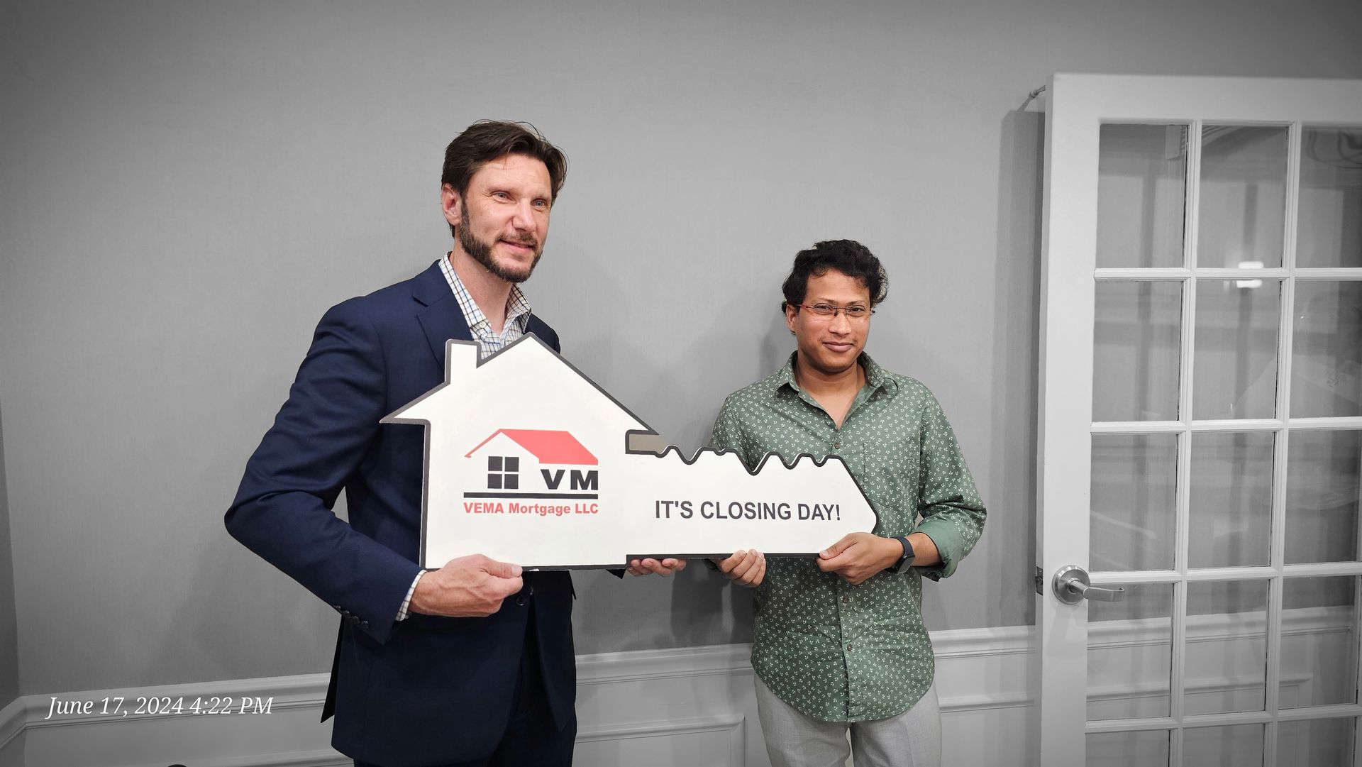 Two people holding a white house-shaped sign in an office, smiling at the camera.